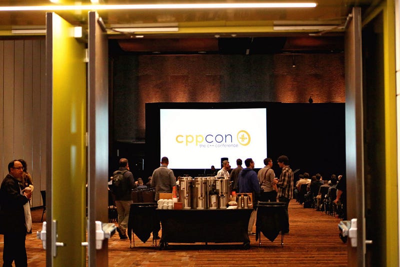 CppCon logo on a screen above attendees in a conference hall